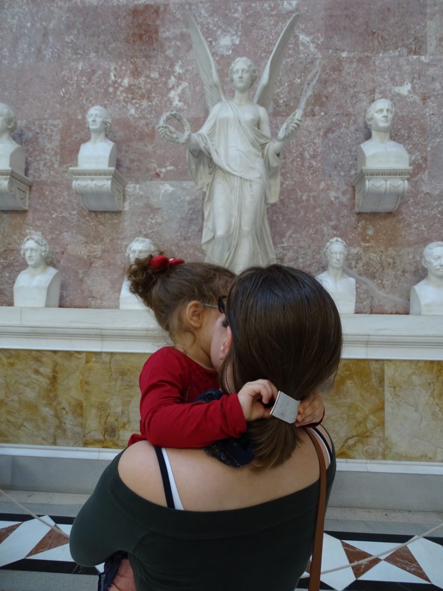 Mama drži ćerku u naručju u dvorani sa mermernim bistama i statuom krilate boginje
