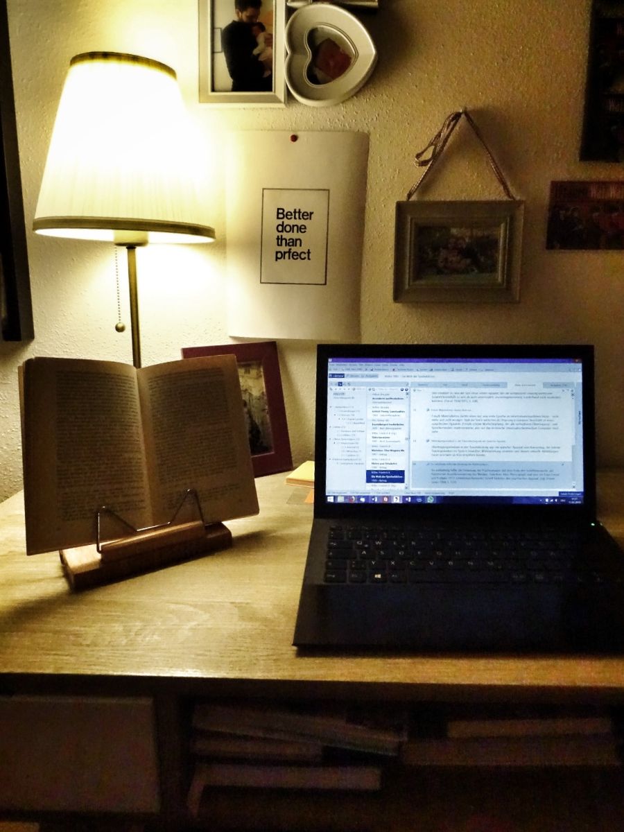A desk in the evening with a laptop, an open book and a lamp while writing a master's thesis