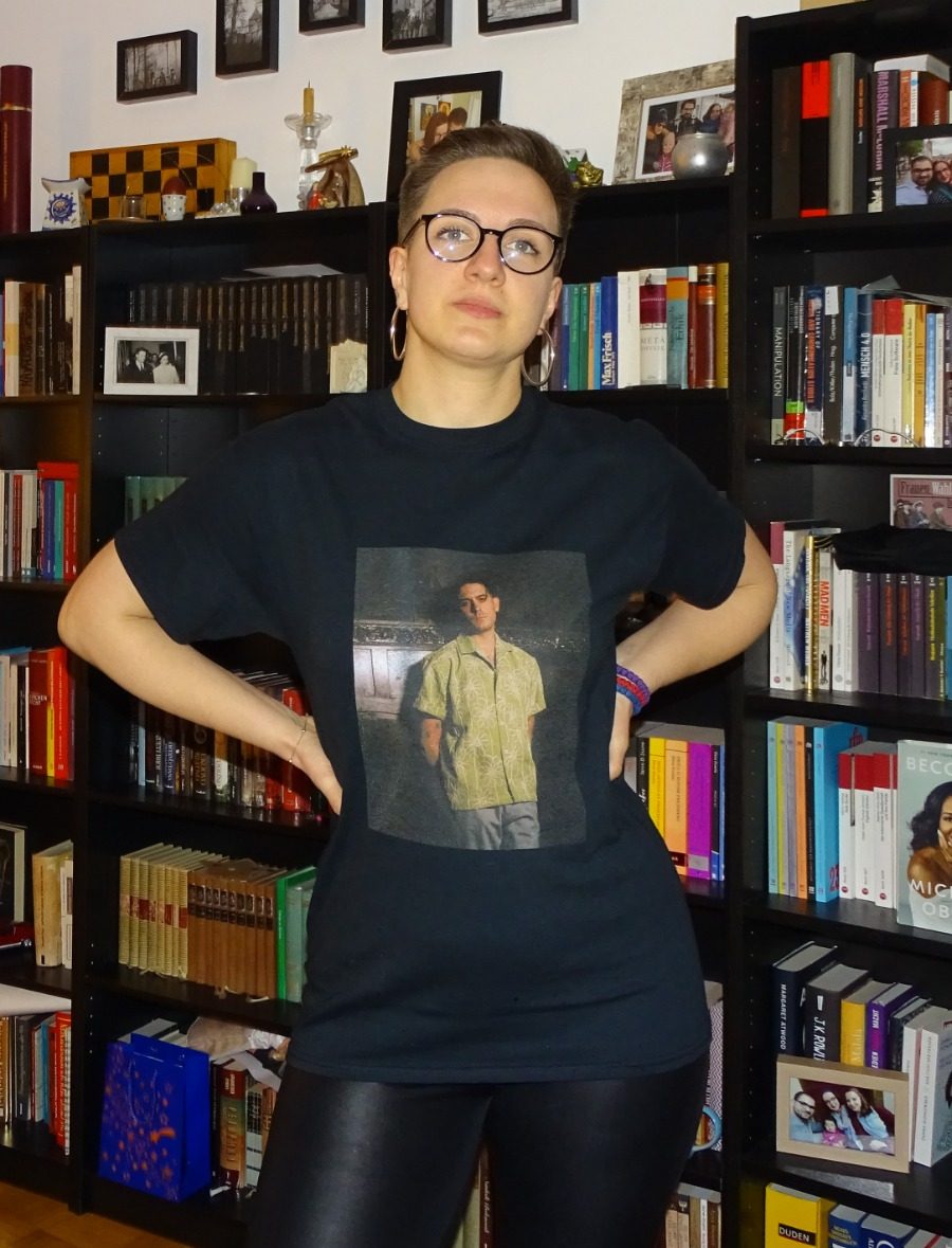 Mima with short hair and glasses, in a black T-shirt and leather leggings, in front of a bookshelf