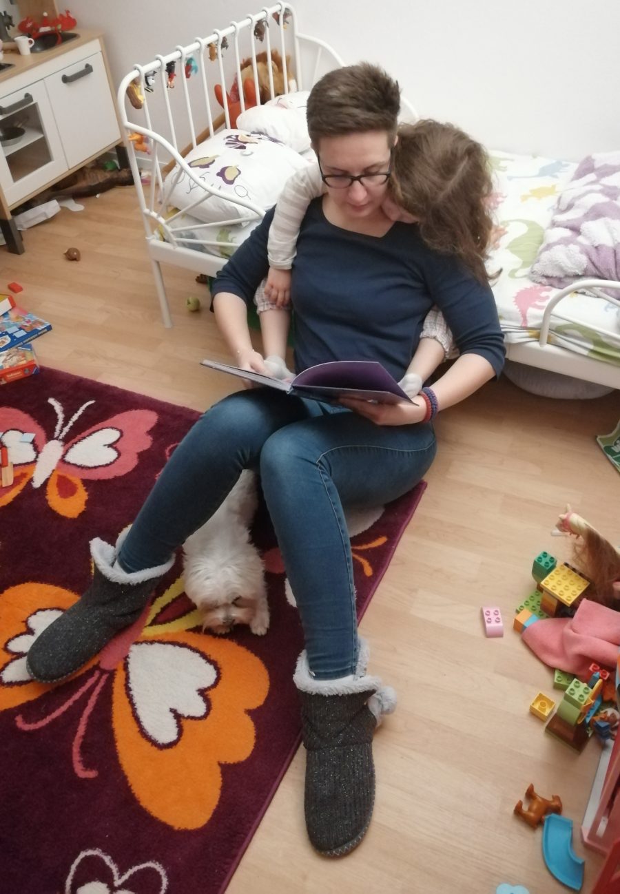 Mama čita knjigu na podu dečje sobe, ćerka joj se privila uz leđa, a pas leži pored nje
